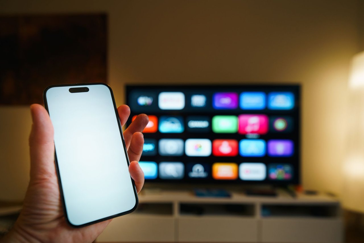Smartphone em primeiro plano diante de uma smart TV com aplicativos exibidos em uma sala de estar.