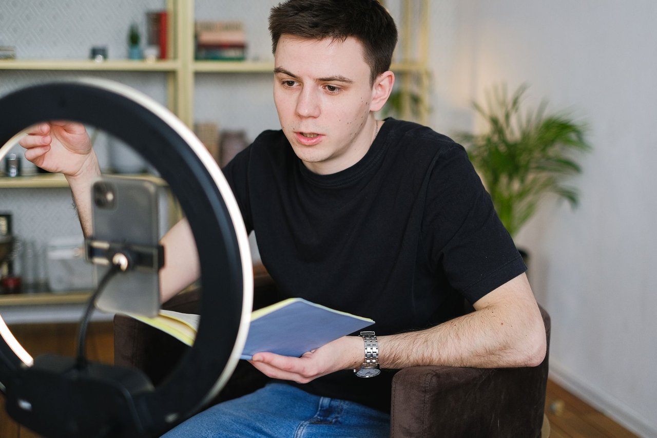 Homem grava conteúdo em ambiente interno usando um smartphone, iluminação circular e segurando um caderno.